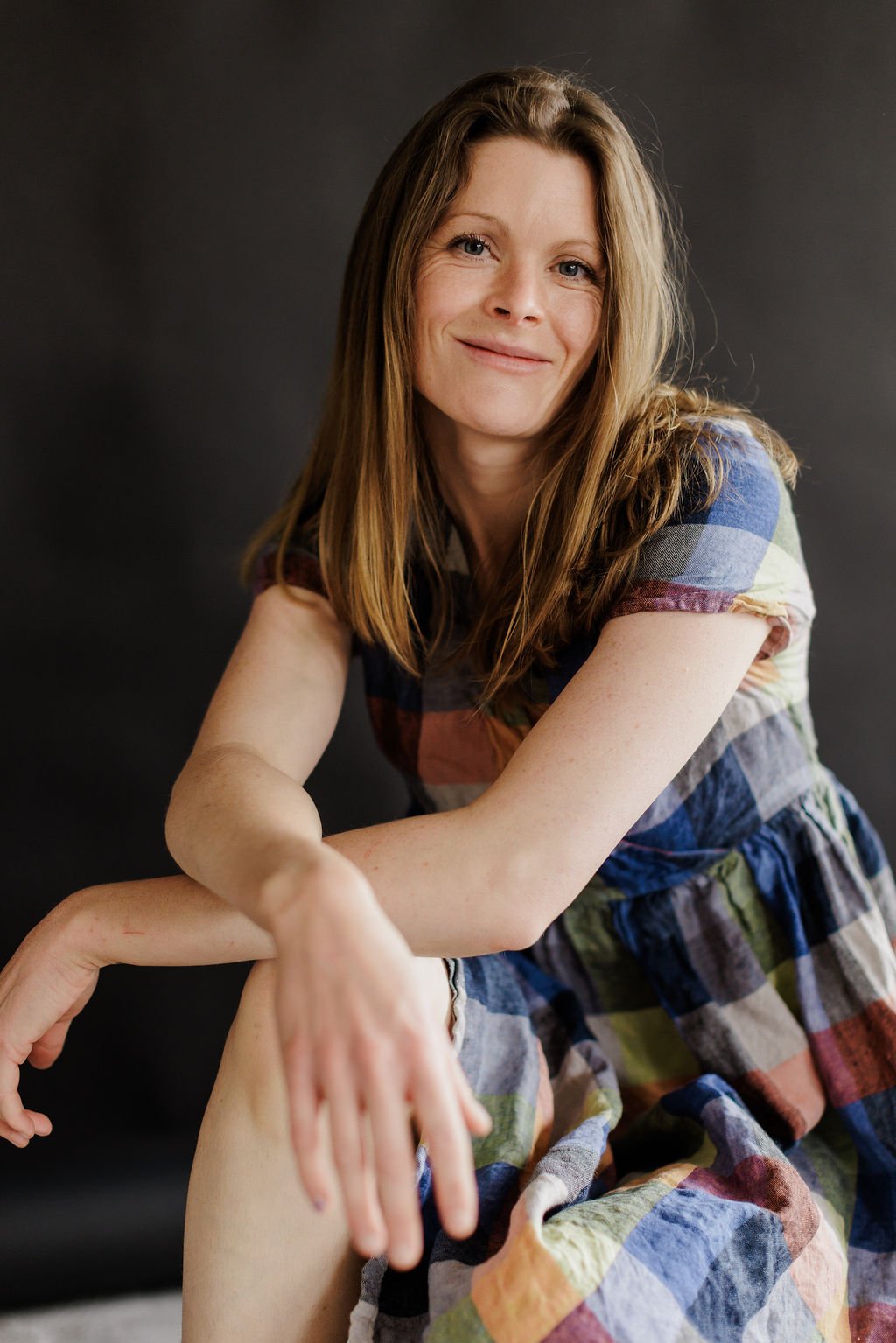Portrait of a smiling woman with long light brown hair, wearing a multicolored plaid dress, against a dark backdrop.